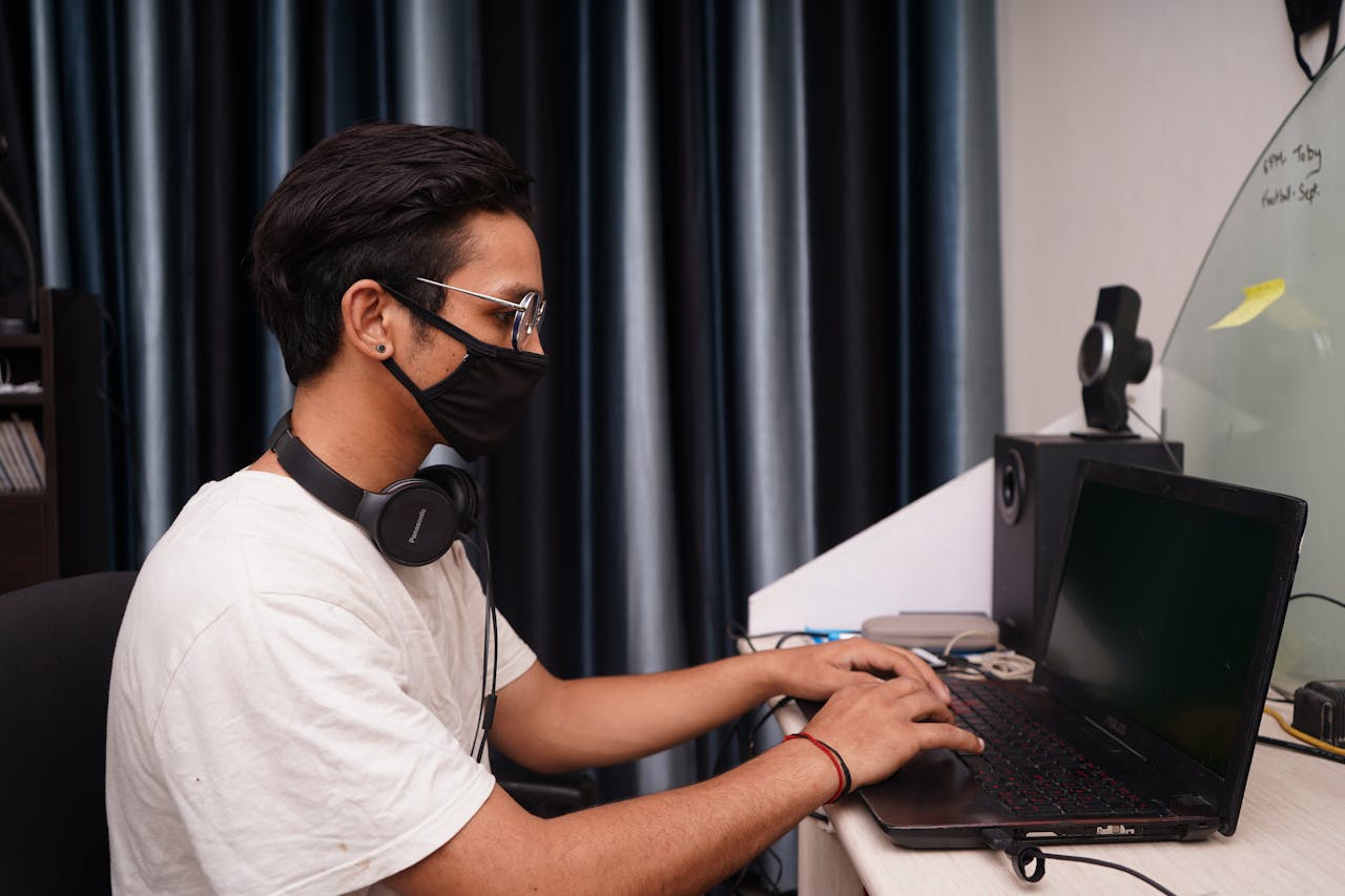 A young man wearing a mask works on his laptop at a home office desk, surrounded by tech accessories.